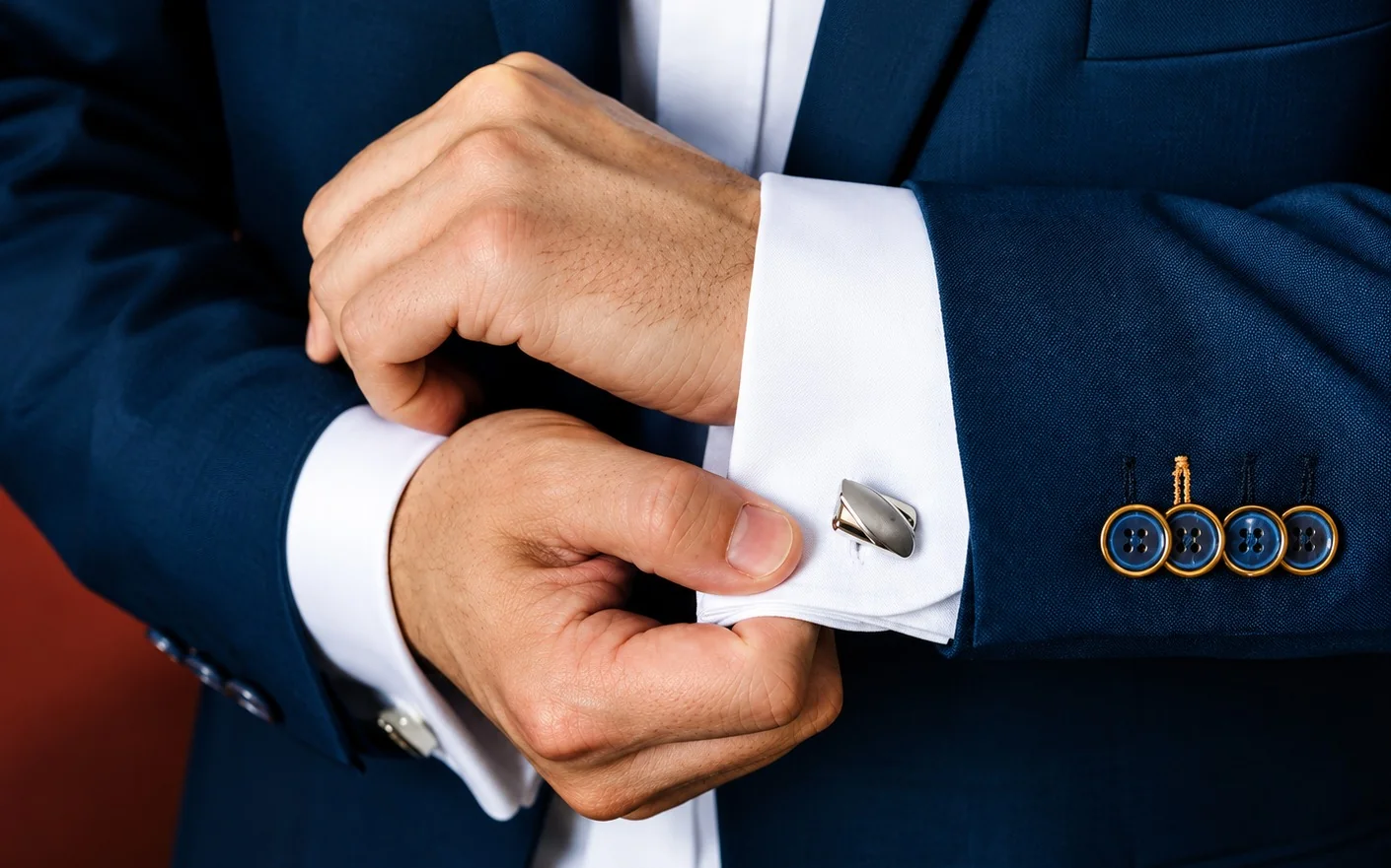 A man in a navy suit adjusts a silver cufflink on the cuff of his white shirt.